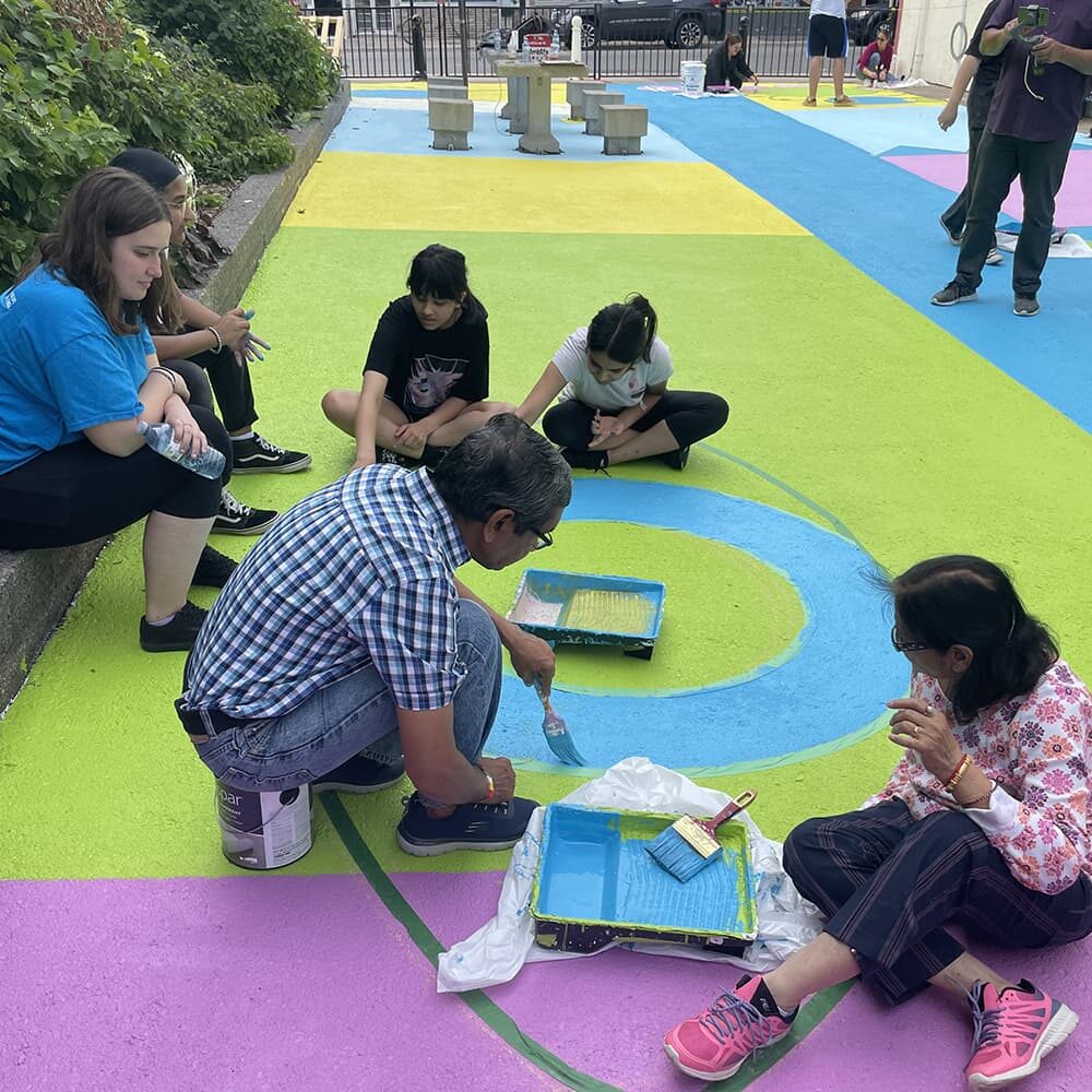 A group of adults and teens painting the floor of a public outdoor space in bright hues of pink, blue and green.