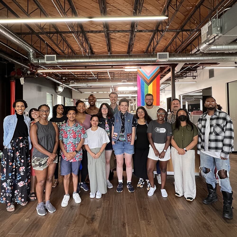 A group photo of twenty young people in an industrial office building with an LGBTQ+ flag hanging in the backgorund.