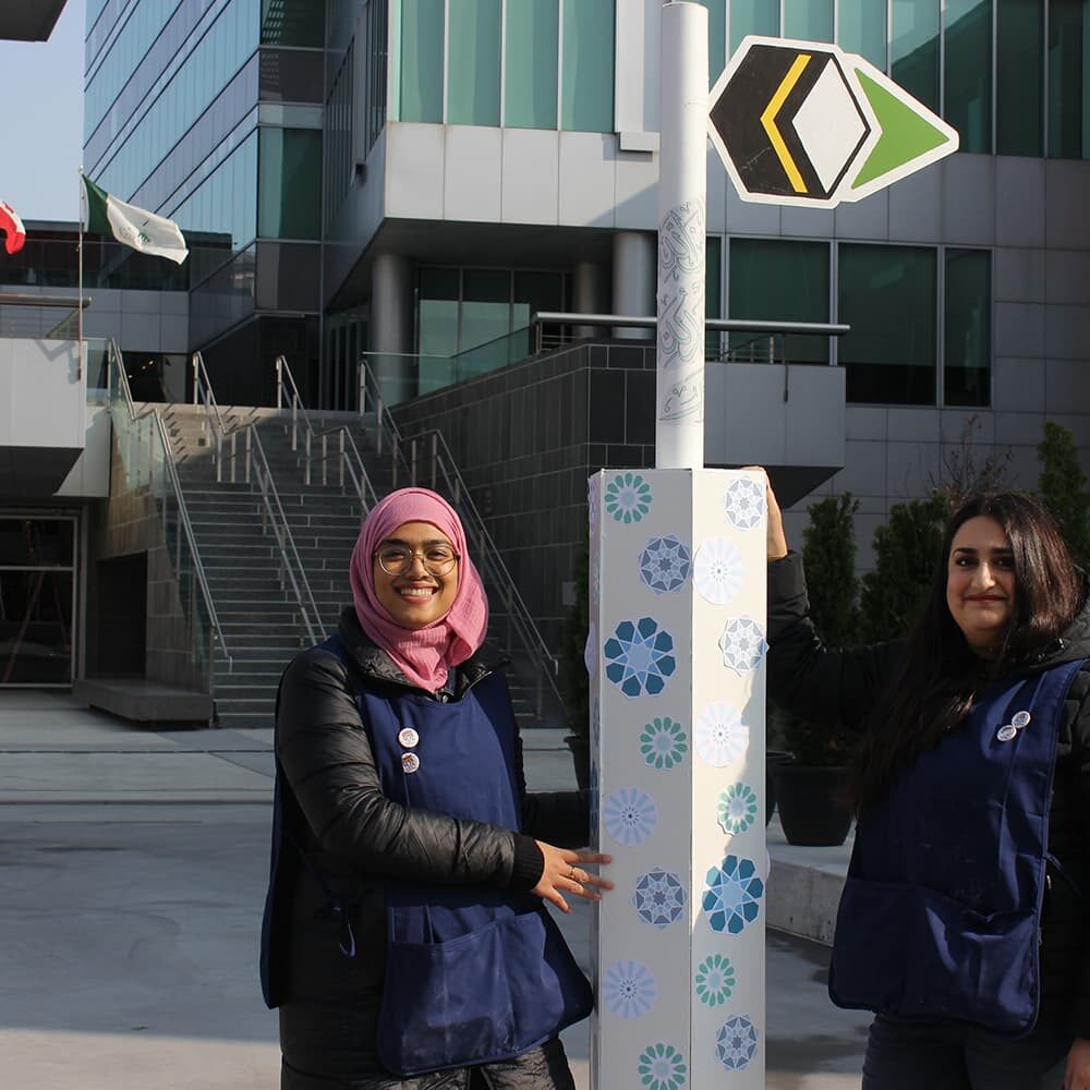 Two young women, one wearing a headscarf, smiling in front of a signage pole.